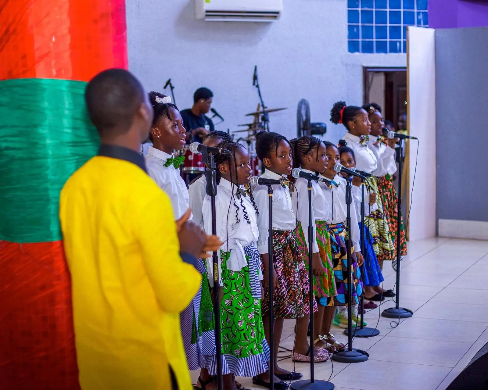 An adult facing a group of pupils singing at microphones